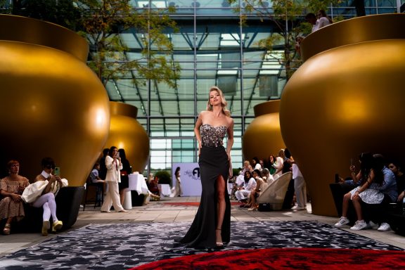 Frau in elegantem Kleid auf einem Laufsteg zwischen großen Goldtöpfen.